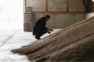 A person kneels in a storage hall next to a large pile of chickpeas, examining them with their hands.
