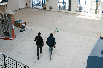 Two people walk through a bright, empty industrial hall or commercial building, photographed from above.